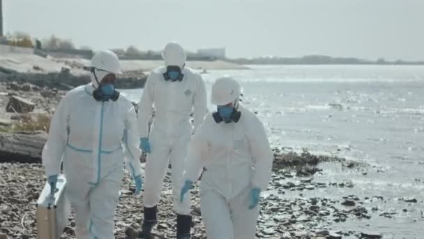 Groep ecologen met beschermende overalls en ademhalingsmaskers die langs de golfkust lopen met steekproefkast en tijdens onderzoek in vervuild gebied discussiëren - Video