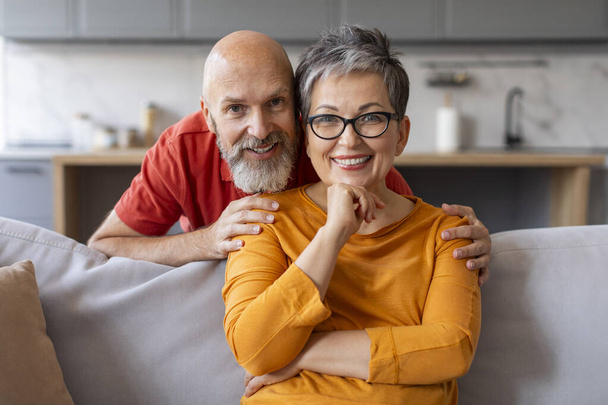 Portrait von glücklichen Senior Ehemann und Ehefrau posiert im Home Interior, liebevolle ältere Ehepartner umarmen und lächeln in die Kamera, entspannen auf der Couch im Wohnzimmer, Mann umarmt Frau von hinten - Foto, Bild