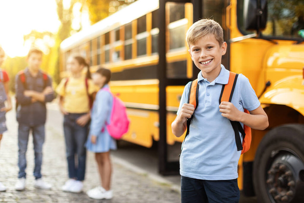 Lächelnder Preteen Boy mit Rucksack posiert neben gelbem Schulbus, während seine Klassenkameraden im Hintergrund plaudern, Nettes männliches Kind bereit für das Studium, Spaß an Bildung und Schulleben, selektiver Fokus - Foto, Bild
