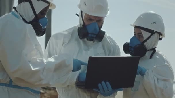 Tým profesionálních ekologů v ochranných kombinézách, rukavicích, helmách a respiračních maskách stojící ve znečištěném prostoru, používajících notebook a mluvící - Záběry, video