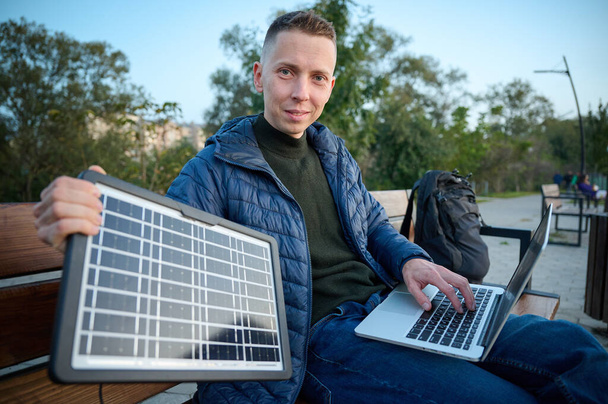 Уверенный бизнесмен сидит на скамейке и заряжает ноутбук портативной солнечной панелью. Концепция возобновляемых источников энергии. Зеленая независимая энергия. - Фото, изображение