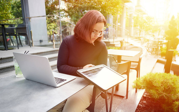 Vrouw die draagbaar zonnepaneel gebruikt om laptop en smartphone op te laden. Portret van een jonge studente die op afstand werkt vanaf een laptop terwijl ze aan een tafel zit in een café. Een stralende zonnige dag. Hernieuwbare energie. - Foto, afbeelding