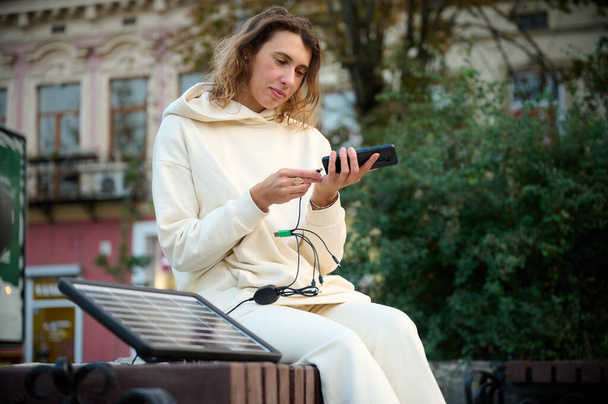 Junge hübsche Frau lädt ihr Handy mit einem tragbaren Solarpanel auf. Solares Batterieladegerät für mobile Geräte. Konzept nachhaltiger Lebensweise und grüner erneuerbarer Energien - Foto, Bild