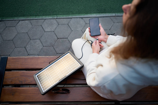 Młoda piękna kobieta ładuje telefon przenośnym panelem słonecznym. Ładowarka baterii słonecznych do urządzeń mobilnych. Pojęcie zrównoważonego stylu życia i ekologicznej energii odnawialnej - Zdjęcie, obraz