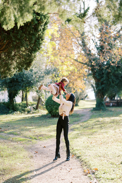 Smiling mother throws up a little girl in her arms while standing on a path in the park. High quality photo - Photo, Image