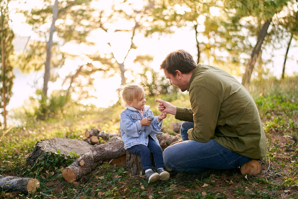 Ο μπαμπάς απλώνει ένα χορτάρι σε ένα χαμογελαστό κορίτσι που κάθεται σε κούτσουρα στο δάσος. Υψηλής ποιότητας φωτογραφία - Φωτογραφία, εικόνα