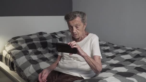 90代のシニア女性は,自宅のベッドに座っているスマートフォンの使い方を学ぶ. 高齢の女性は,退職後の家庭で携帯電話を使用する方法について教育を受けます. テクノロジーを使った老婦人研究.  - 映像、動画