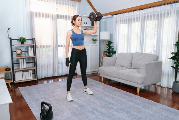 Krachtige energieke vrouw doet halter gewicht tillen oefening thuis. Jonge atletische aziatische vrouw kracht en uithoudingsvermogen training sessie als thuis workout routine. - Foto, afbeelding
