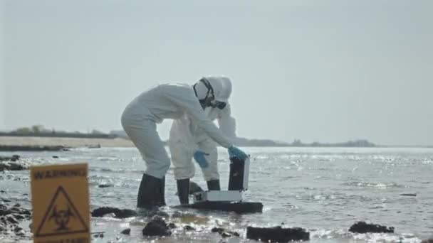Два эколога в защитных комбинезонах и респираторных масках собирают образцы воды из озера на загрязненной территории с предупреждающим знаком биологической опасности - Кадры, видео