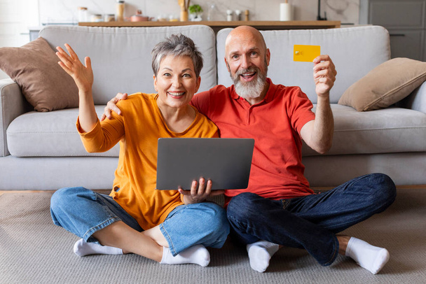 Koncepcja bankowości elektronicznej. Podekscytowani starsi małżonkowie z laptopem i kartą kredytową pozujących we wnętrzu domu, szczęśliwa para w podeszłym wieku korzystająca z płatności online i łatwych przelewów pieniężnych, uśmiechnięta przed kamerą - Zdjęcie, obraz