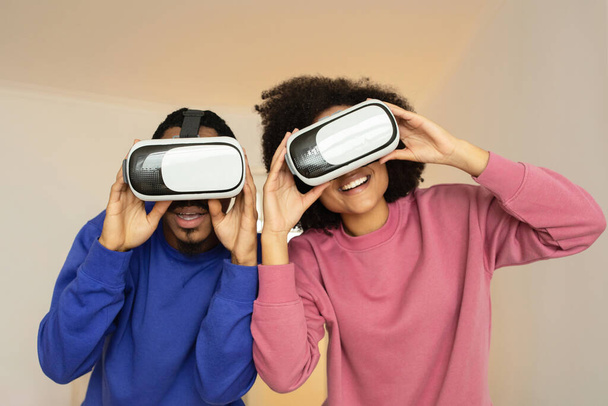 VRメガネを着てポーズするアフリカ系アメリカ人のカップルは,未来的なゲームセッションを楽しんで,ゴーグルを通してカメラを見ながらスタジオウォールに対して遊び心を持ってポーズします. 拡張されたデジタル楽しい - 写真・画像