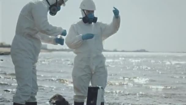 Dos ecologistas en trajes de protección y máscaras respiratorias recogiendo muestras de agua del lago en área contaminada en tubo de ensayo - Metraje, vídeo