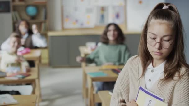 落ち込んだオタクの少女の選択的なフォーカスミディアムクローズアップ肖像画は,教室に立っているコピーブックを保持し,彼女のクラスメートは彼女に紙のボールを投げます - 映像、動画