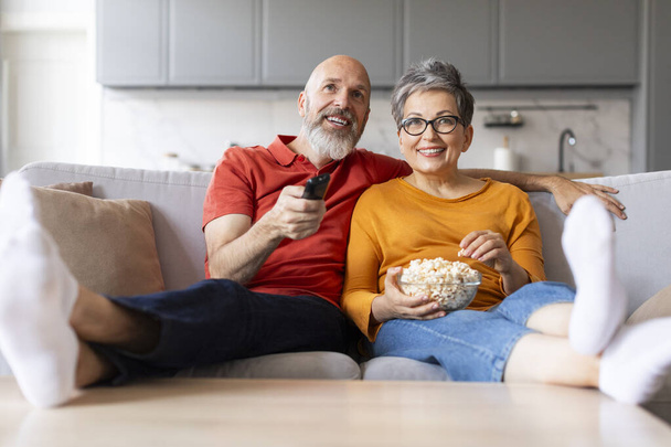Портрет щасливої заміжньої старшої пари Дивитися телевізор і їсти попкорн вдома, усміхнений чоловік і дружина приймає і розслабляється на зручному дивані у вітальні, чоловік тримає пульт дистанційного керування - Фото, зображення