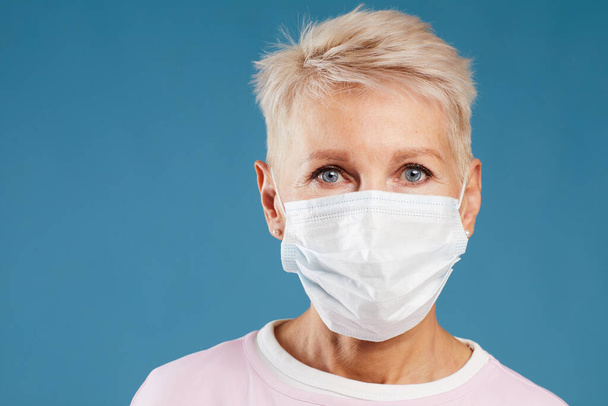 Portrait of blond-haired female doctor in medical mask for protection of infections standing against blue background - Foto, Imagen