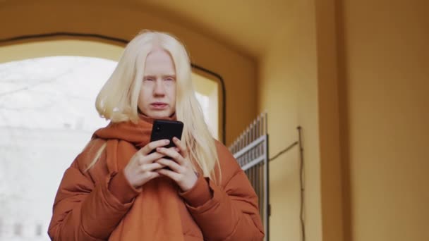 Alacsony szögű lassított felvétel portré fiatal nő albínizmus visel stílusos meleg ruhát áll a szabadban ellenőrzése valamit okostelefon - Felvétel, videó