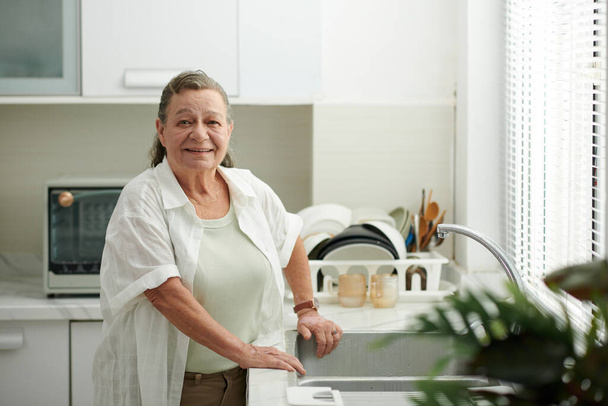Retrato de mujer mayor sonriente de pie en el fregadero de la cocina - Foto, imagen
