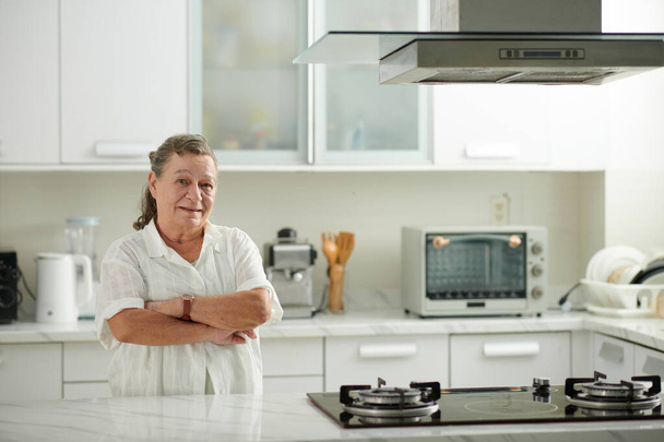 Mosolygó idős nő portréja, amint a konyhaasztalnál áll, karokkal keresztbe tett kézzel, miután letisztította a felületeket - Fotó, kép