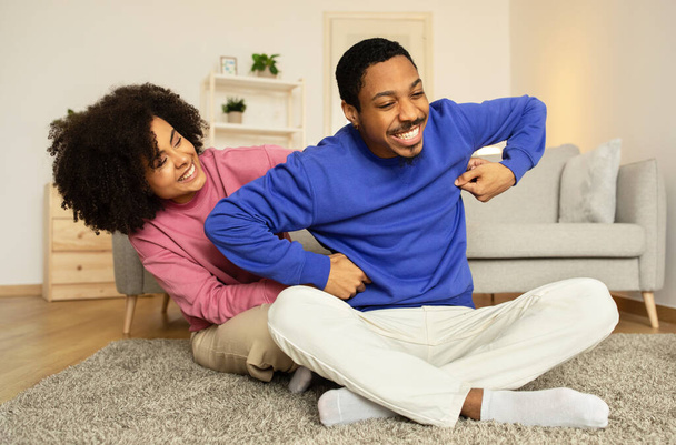 Cheerful black wife bonding with husband through gentle tickles, laughing in joy and affection, having fun on lazy weekend in modern home interior, sitting on floor together. - Foto, immagini