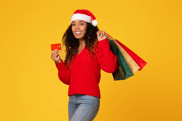 Veselá černoška v klobouku Santa drží kreditní karty a nákupní tašky, připravena k prodeji na dovolenou, stojí na žlutém pozadí a usmívá se na kameru - Fotografie, Obrázek