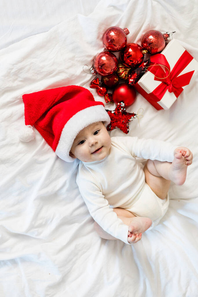 Natale bambino in cappello Babbo Natale seduto sul letto con scatola regalo e bagattelle festive. - Foto, immagini