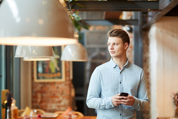 Serious pensive young manager in casual shirt standing in contemporary cafe and looking away thoughtfully while texting sms on phone - Foto, imagen
