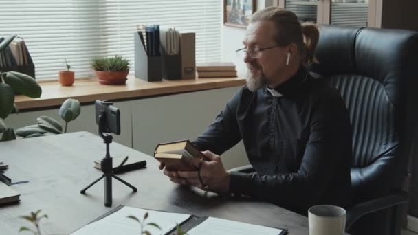 Modern mature Catholic priest wearing earbuds sitting at desk in office recording video on smartphone for his blog - Footage, Video
