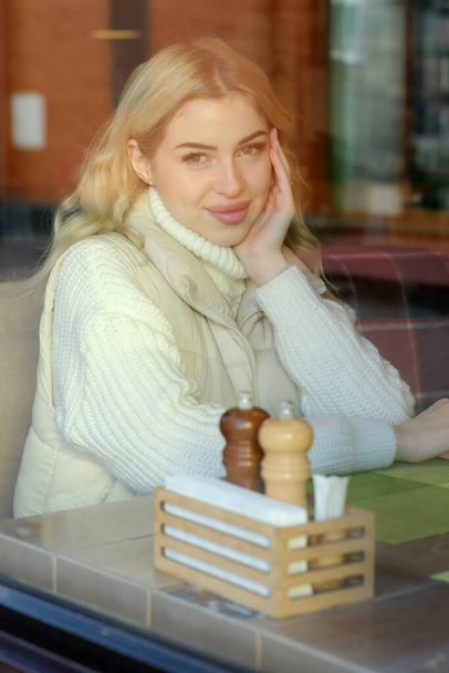 Retrato de una joven rubia hermosa con el pelo largo en un suéter blanco y chaqueta descansando en un café, vista desde la calle a través de la ventana - Foto, imagen