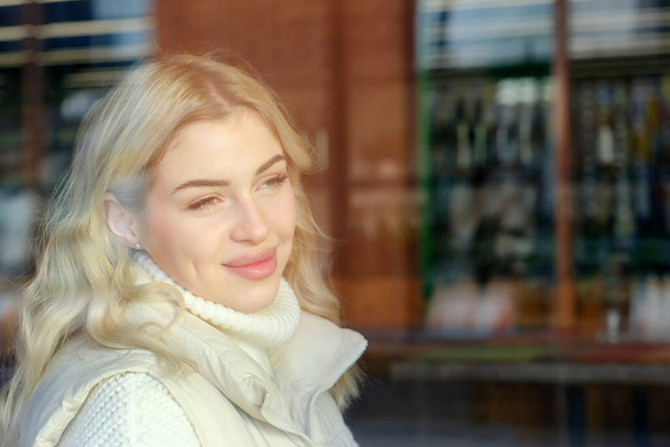 Portrait of a young beautiful blonde with long hair in a white sweater and jacket resting in a cafe, view from the street through the window - Photo, Image