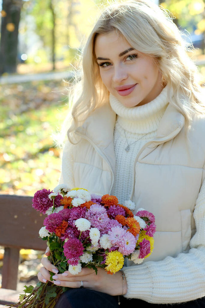 Portré egy fiatal, gyönyörű szőke, hosszú hajú, fehér pulóverben és dzsekiben napfényes ősszel egy városi parkban egy csokor virággal - Fotó, kép