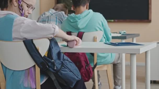 Белая школьница-подросток достает канцелярские принадлежности из сумки, сидя за столом во время урока. Женщина-учитель объясняет тему на доске в размытом фоне - Кадры, видео