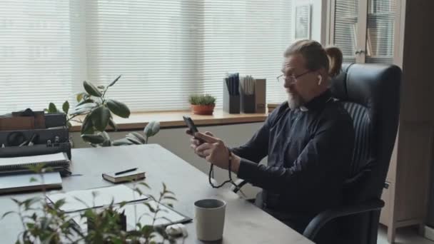 成熟した白人カトリック司祭は,ビデオ通話で話している彼のオフィスに座ってワイヤレスイヤホンを身に着けています - 映像、動画