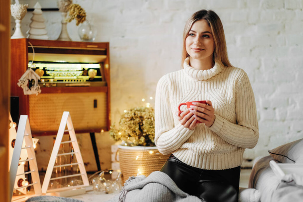 Μια νεαρή όμορφη γυναίκα κάθεται σε ένα σπίτι κοντά στο παράθυρο την παραμονή των Χριστουγέννων με μια κούπα ζεστό τσάι. - Φωτογραφία, εικόνα