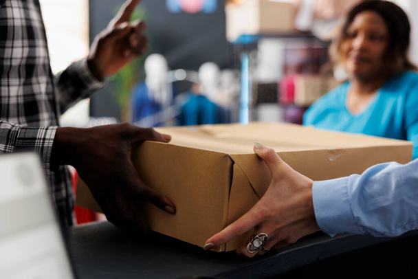 Shopaholic man holding cardboard box at counter desk, after buying fashionable merchandise. African american man shopping for stylish clothes, discussing about material in modern boutique - 写真・画像