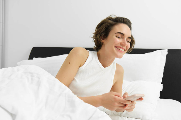 Πορτρέτο της γυναίκας στο κρεβάτι, ξαπλωμένη σε λευκά σεντόνια, χρησιμοποιώντας smartphone, κρατώντας το κινητό τηλέφωνο. Έννοια του τρόπου ζωής και τεχνολογίας - Φωτογραφία, εικόνα