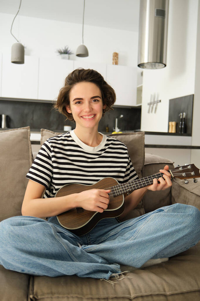 Pionowe ujęcie młodego muzyka, kobieta uczy się grać na ukulele, siedzi na kanapie ze skrzyżowanymi nogami i uśmiecha się do kamery. - Zdjęcie, obraz