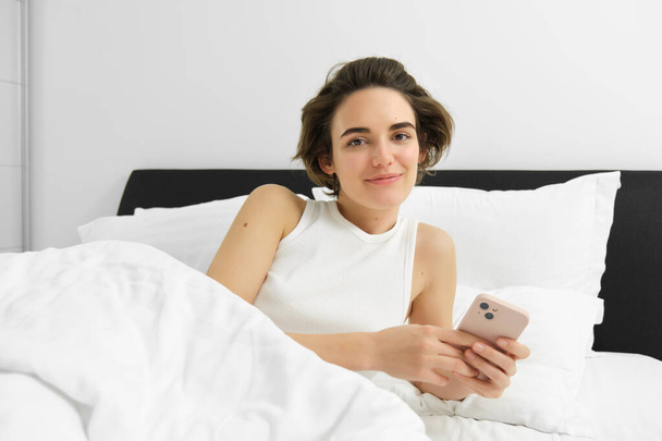 Genç bir kadının portresi yatakta uyanır, akıllı telefon kullanır, cep telefonunun alarmını kapatır, yatak odasında beyaz çarşafların üzerinde yatar.. - Fotoğraf, Görsel
