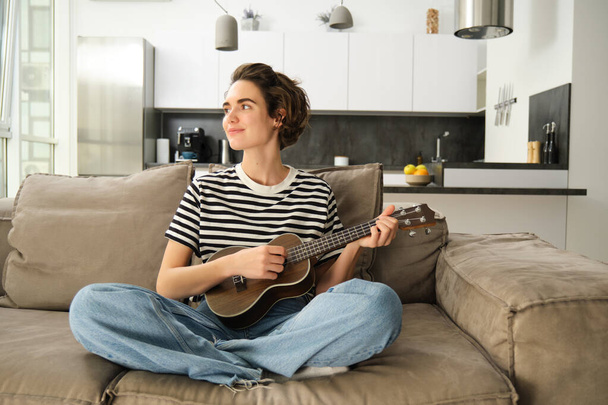 Ritratto di ragazza che riposa in salotto, suona l'ukulele, impara a suonare nuovo strumento musicale, trascorrendo del tempo a rilassarsi a casa. - Foto, immagini
