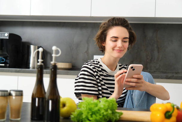 Portrait de jeune femme à la recherche de recettes de cuisine en ligne à l'aide d'un smartphone, assise près de légumes, ingrédients de salade et planche à découper, souriant à la caméra. - Photo, image