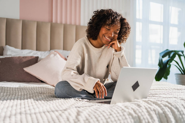 Chica afroamericana que usa el ordenador portátil en la cama en la oficina de casa escribiendo chateando leyendo escribiendo correo electrónico. Joven mujer negra que tiene una reunión virtual de chat en línea de videoconferencia. Aprendizaje laboral desde casa - Foto, imagen