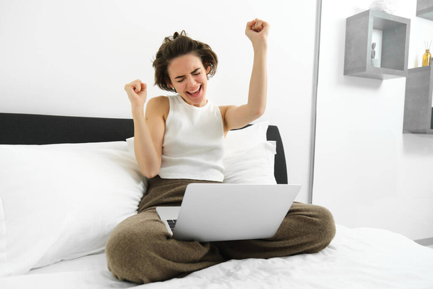 Портрет счастливой женщины, радующейся, сидящей за компьютером и танцующей победительницу, с ноутбуком на коленях, поднимающей руки и празднующей, торжествующей от радости. - Фото, изображение