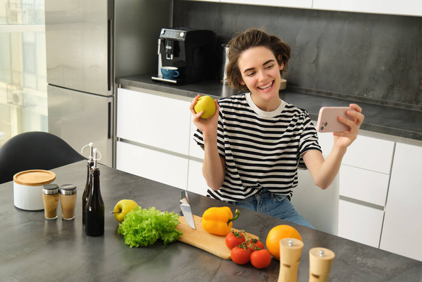Smiling young woman, food blogger recording a video on smartphone with food, cooking and making selfie, taking pictures during meal preparation, sitting near vegetables and holding an apple. - 写真・画像