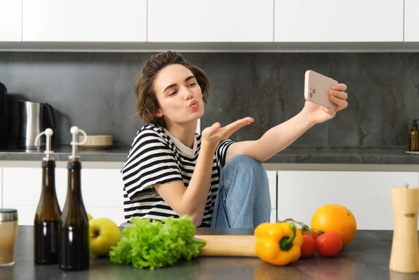 Nette junge Frau, Lifestylebloggerin beim Kochen, mit Gemüse und Schneidebrett in der Küche sitzen, Selfie mit ihrem neuen Smartphone machen, Küsse vor der Handykamera verschicken. - Foto, Bild