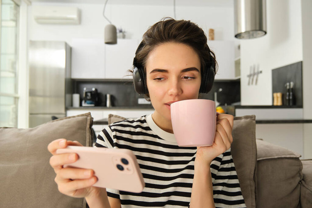 Portrait of woman in her living room, sitting on sofa and watching videos on mobile phone, wearing headphones, drinking tea or coffee in pink cup. - Photo, Image