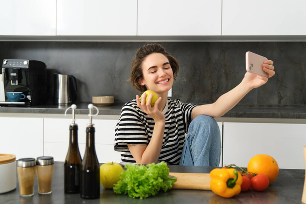 Aranyos fiatal nő, modern életmód blogger, selfie főzés közben saláta a konyhában, okostelefon segítségével fotók a közösségi média app. - Fotó, kép