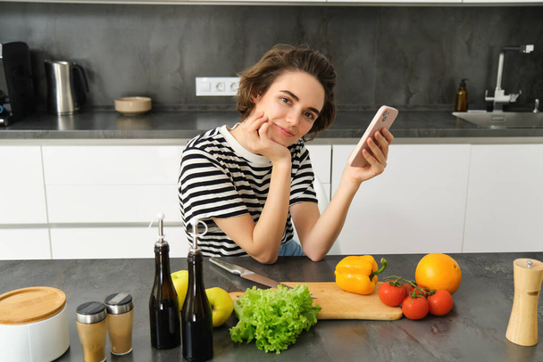 Portrait of cute brunette woman in the kitchen, cooking, searching recipe on smartphone, taking pictures for her food blog, cooking meal from vegetables, vegan salad. - Photo, Image