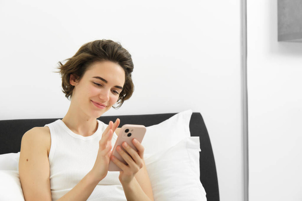 Портрет стильной, современной молодой женщины дома, сидящей на кровати и пользующейся мобильным телефоном, читающей на смартфоне, прокручивающей приложение социальных сетей в спальне. - Фото, изображение