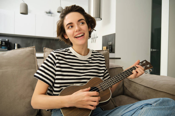 Beautiful young woman playing ukulele, singing and enjoying her musical instrument, spending time alone at home, relaxing in living room. - Photo, Image