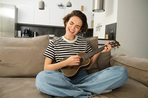 Portrait of cheerful young woman playing her ukulele, singing and laughing, sitting in living room at home. Lifestyle and music concept - Photo, Image