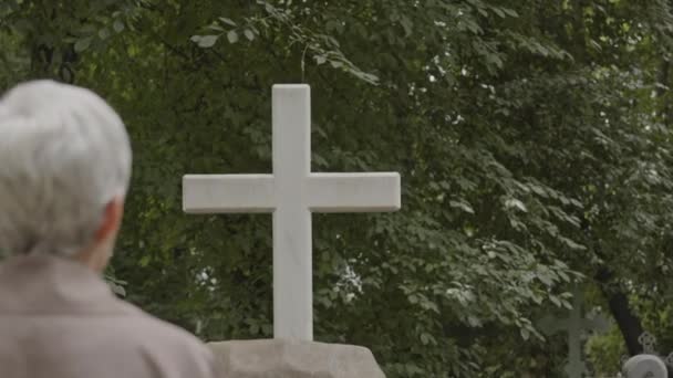 Widok na tył starej wdowy patrząc na granitowy pomnik krzyża podczas jej wizyty na cmentarzu w godzinach porannych - Materiał filmowy, wideo